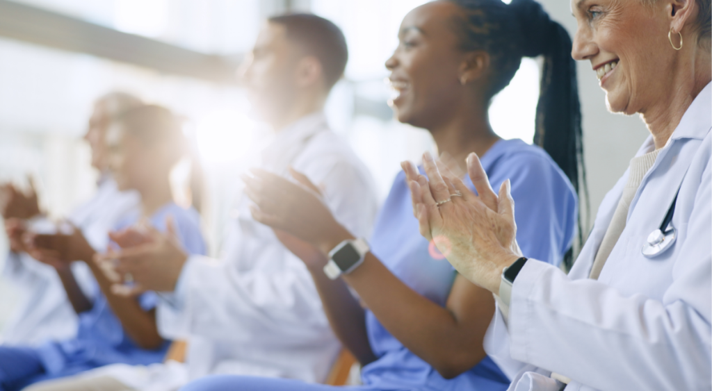 A diverse group of healthcare professionals in white coats and blue scrubs sitting in a row and smiling while they applaud.