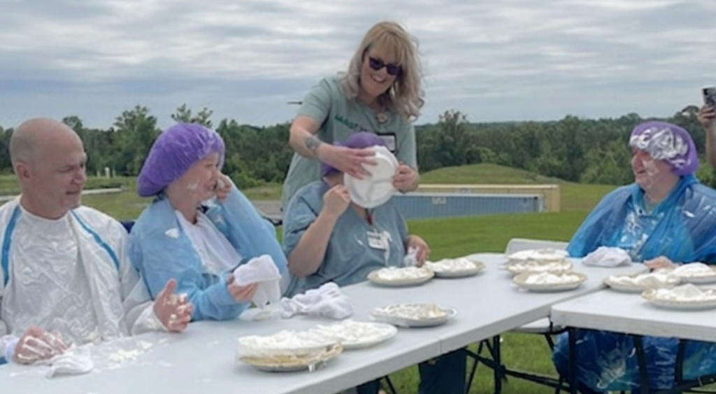 An outdoor pie-eating contest where participants sit at a long white table covered in whipped cream, wearing protective ponchos and shower caps, while an organizer playfully presses a pie into a participant's face.
